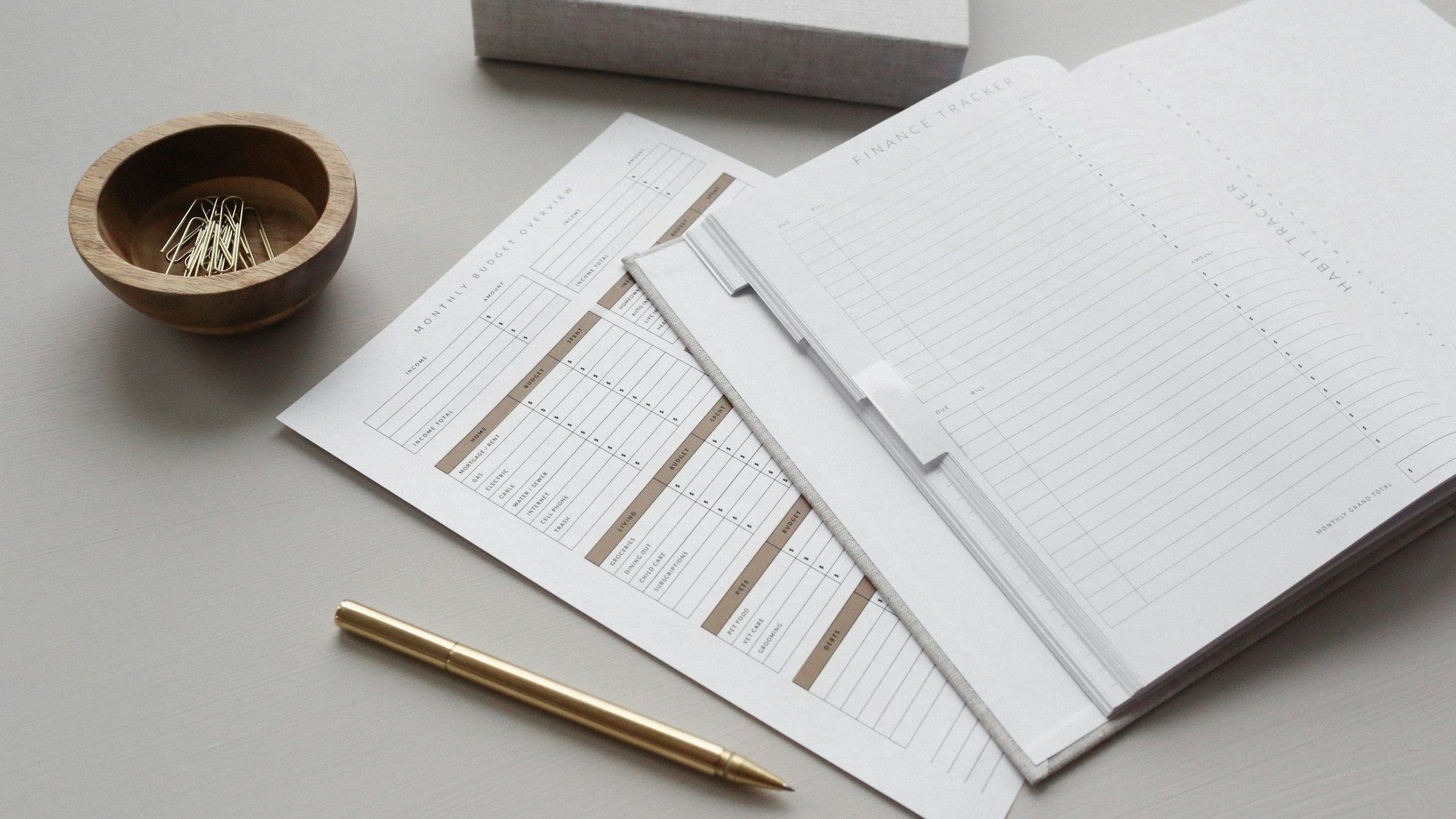 Several printed sheets with tables and a white folder on a pale table, with a gold pen and a bowl of paper clips