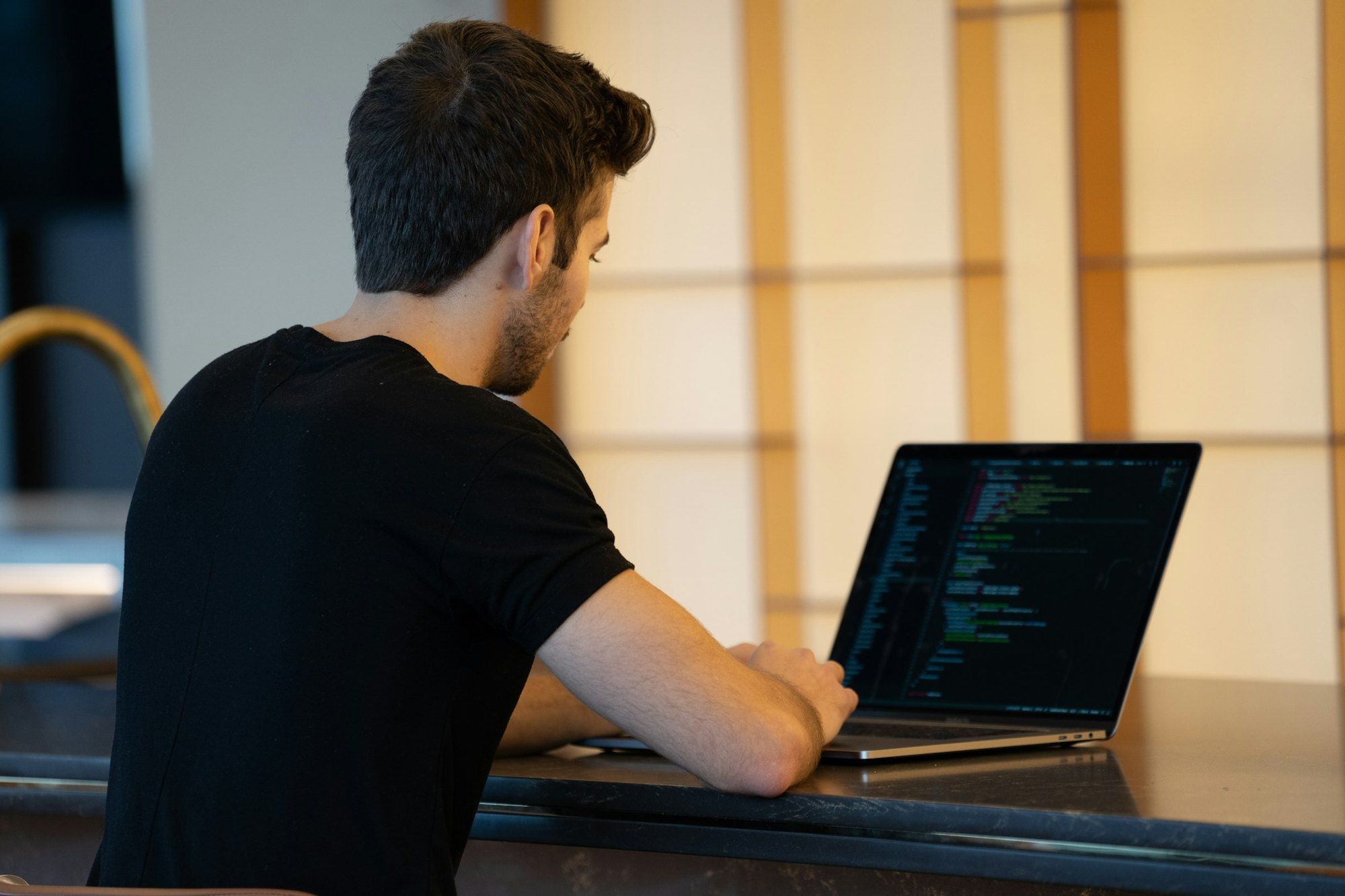 A man seen from behind, in a black T-shirt, works with code on a laptop in front of a translucent panel of warm light