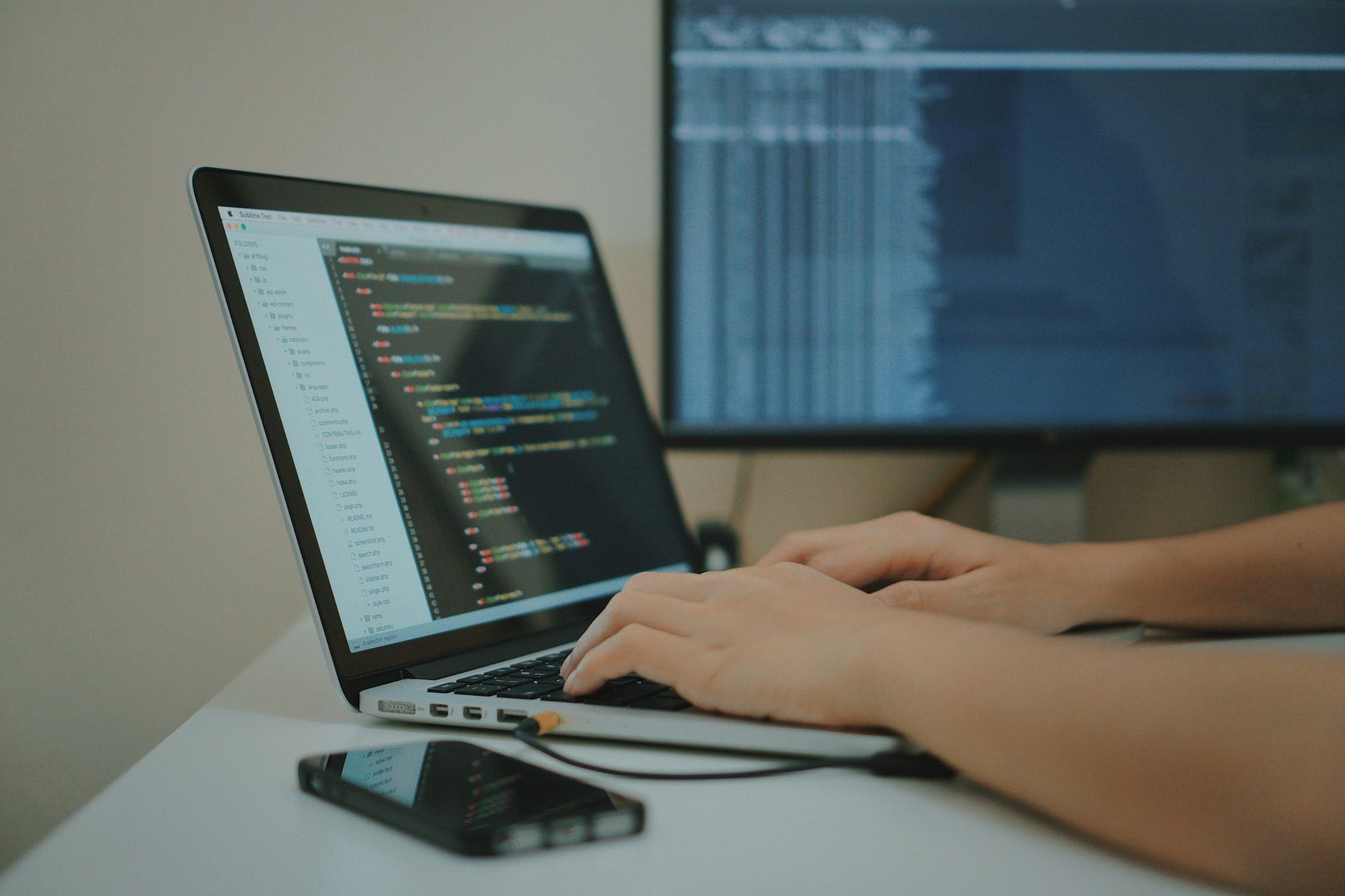 Hands typing on a laptop showing a code editor, with a large monitor behind and a phone on the white desk