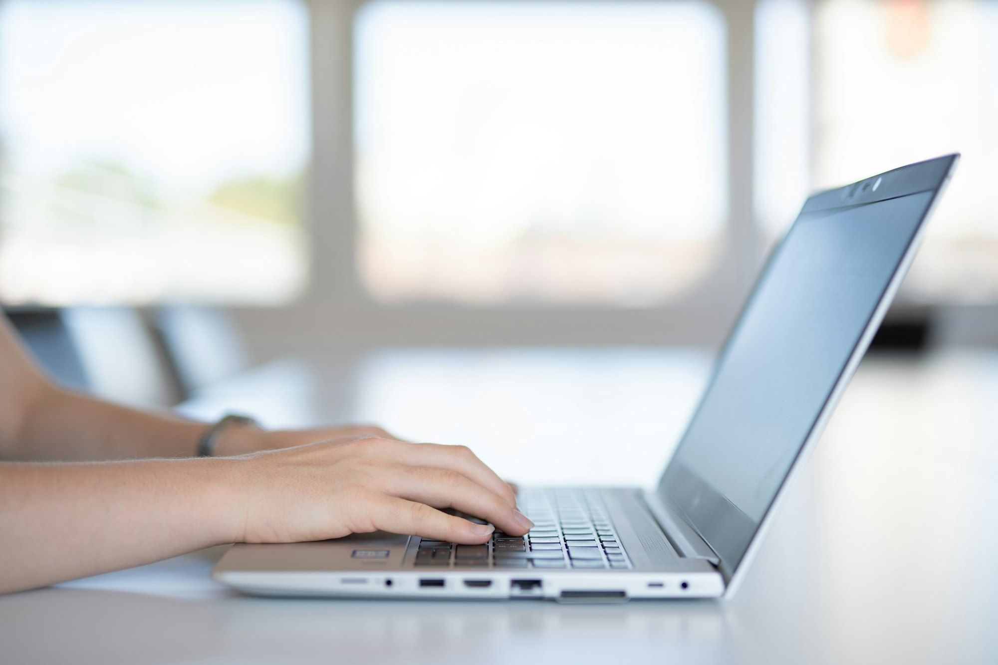 Hands typing on a silver laptop on a white table, with a bright window out of focus behind