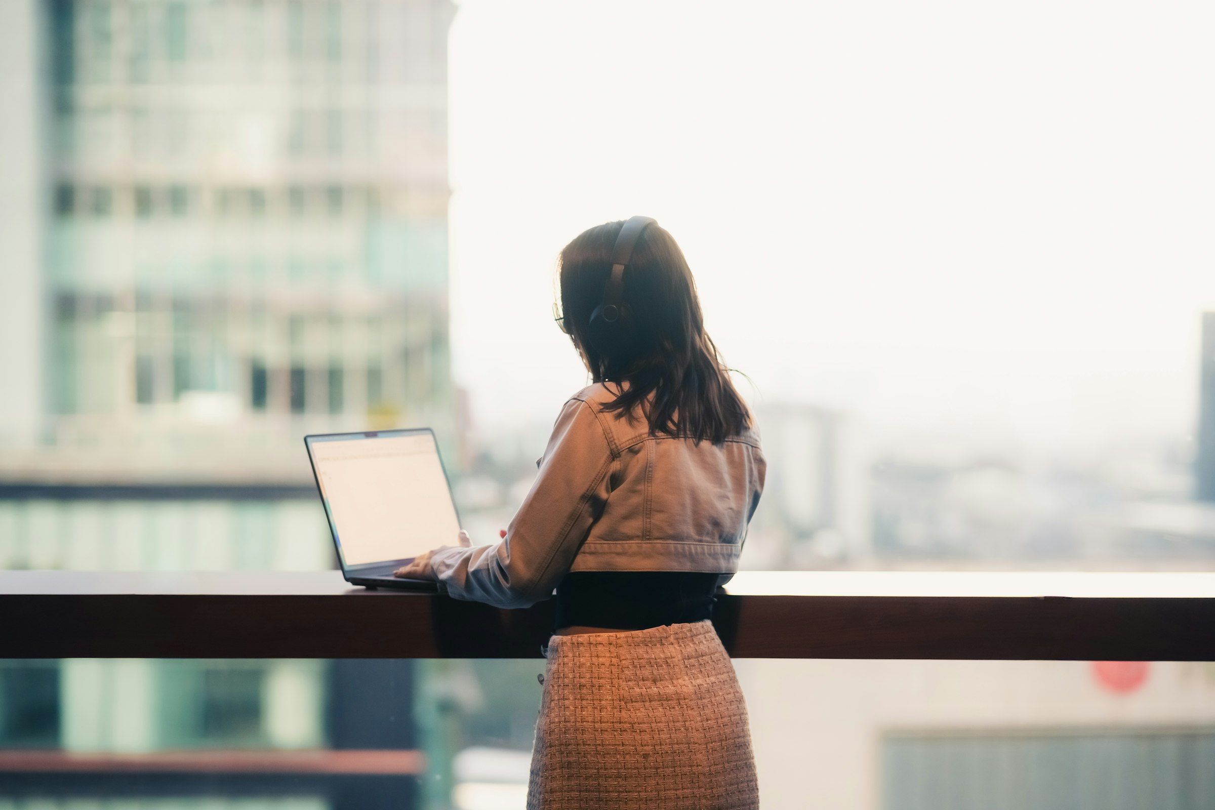 A woman seen from behind, wearing headphones, works on a laptop resting on a counter in front of a large window with the city blurred beyond