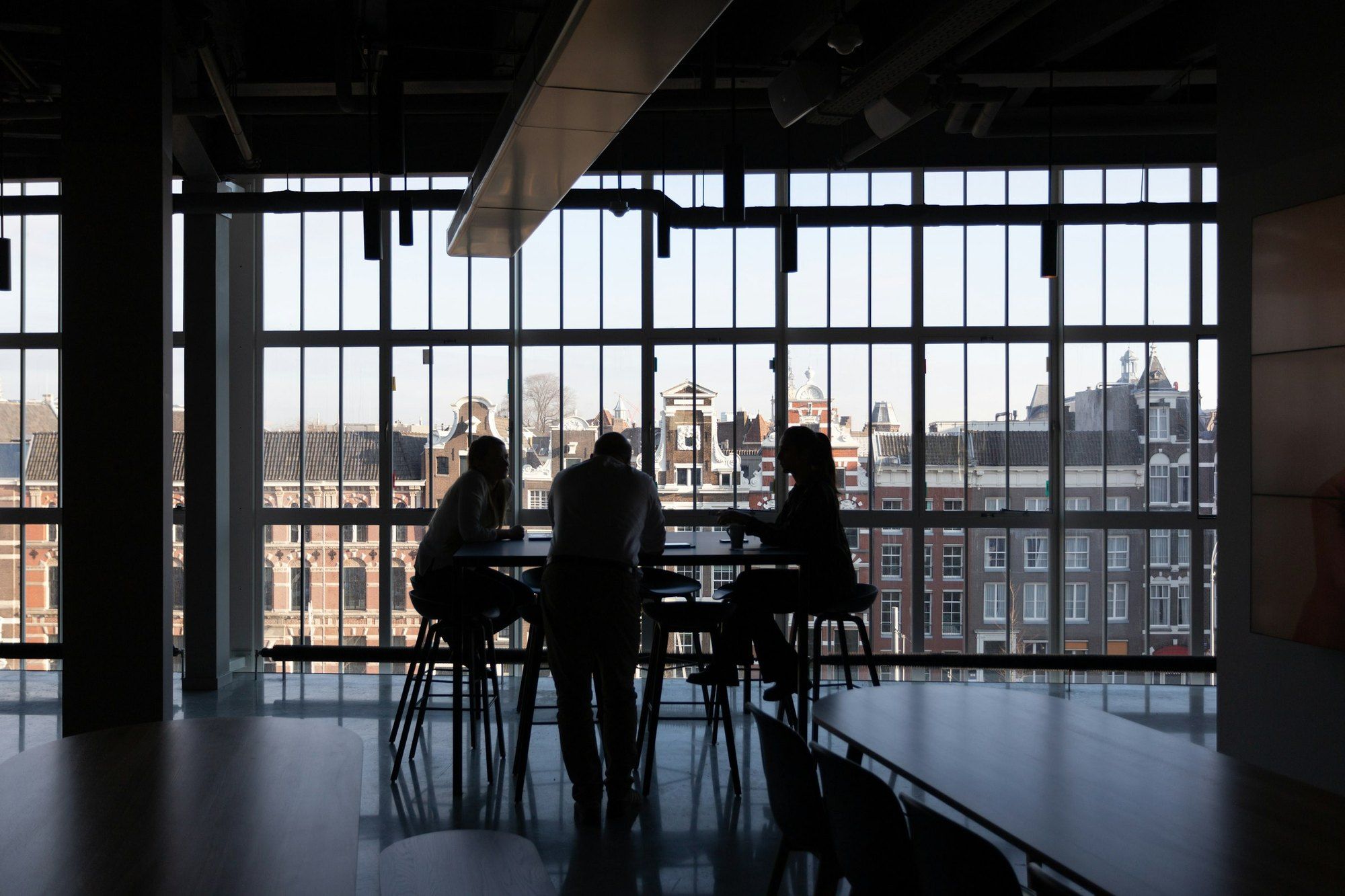 Three people in silhouette talk beside a high table in front of a large window with buildings beyond
