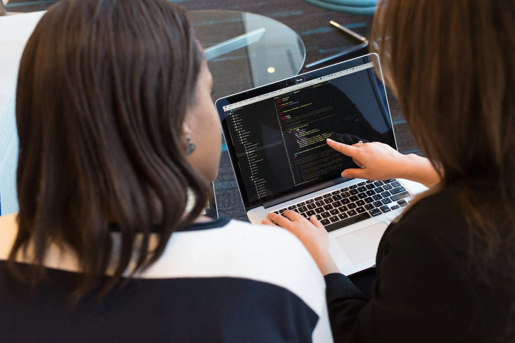 Two people seen from behind review code on a laptop; one points at a line on the screen